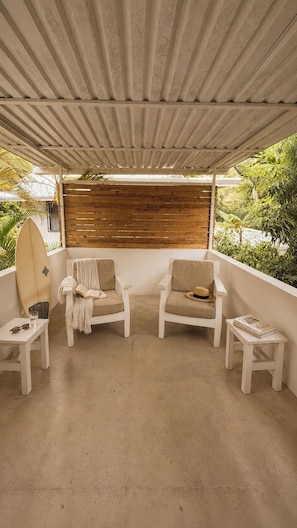 Een overdekt terras met twee witte stoelen, een surfplank en een kleine tafel met een boek en brillen.
