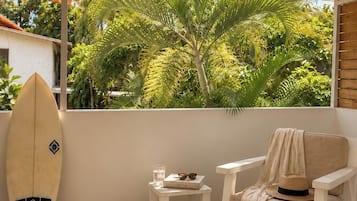 A room with a white ceiling, a window overlooking a palm tree, a white chair, a small table with a book and glasses, and a surfboard leaning against the wall.