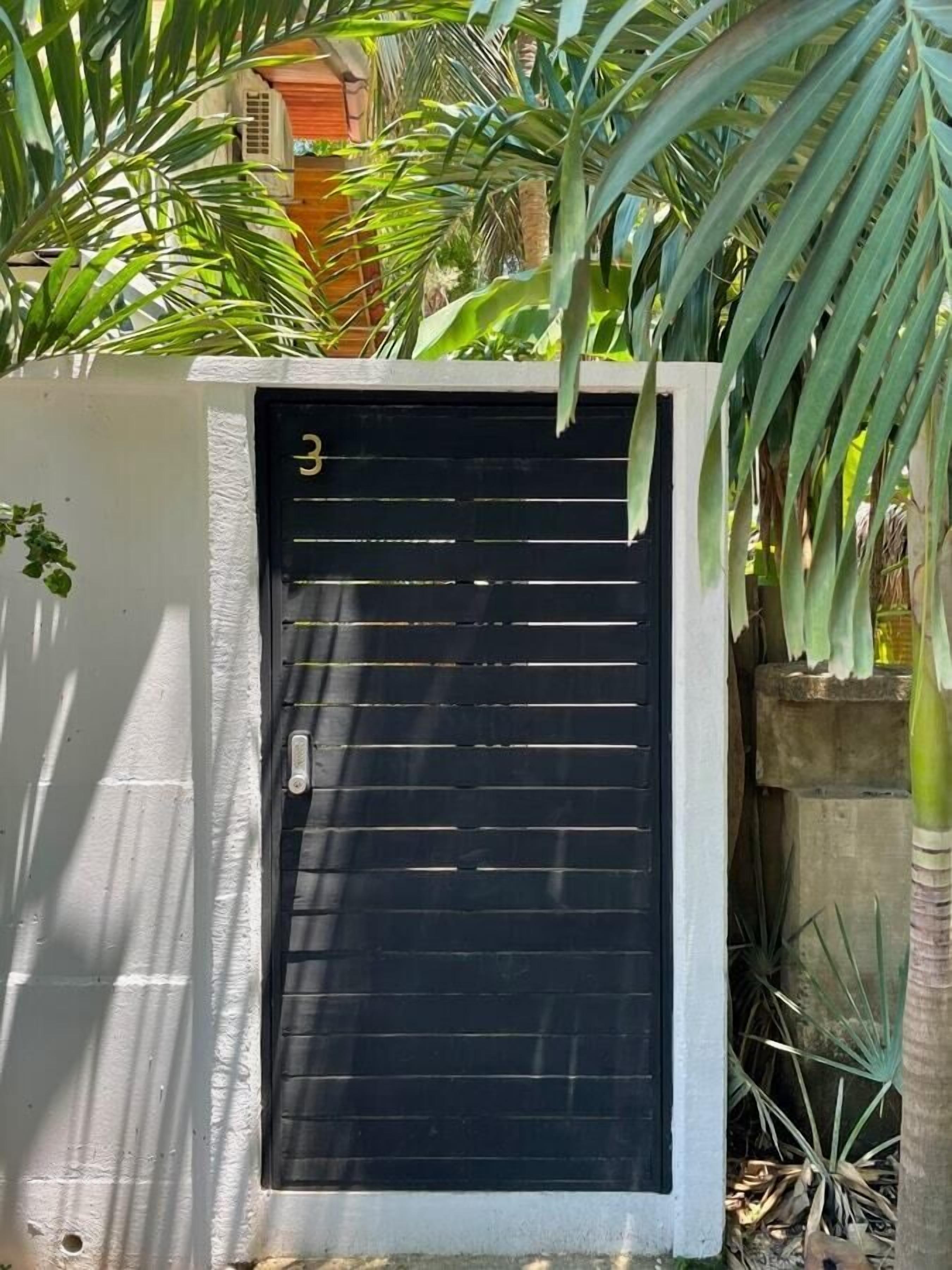 A black gate with the number 3 on a white wall, surrounded by greenery.