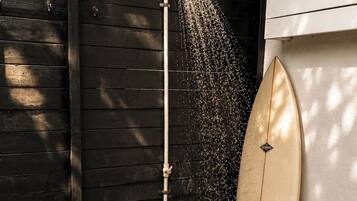 A showerhead with water flowing, a surfboard leaning against a wall, and a garden with plants.