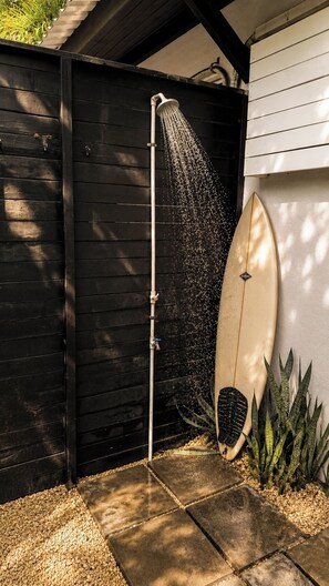 Un cabezal de ducha con agua corriendo, una tabla de surf apoyada contra una pared y un jardín con plantas.
