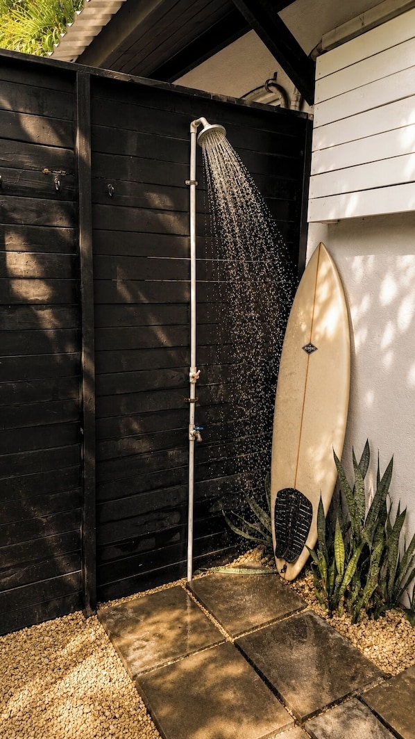 Eine Duschbrause, darunter fließt Wasser, ein Surfbrett, das an eine Wand gelehnt ist, und ein Garten mit Pflanzen.
