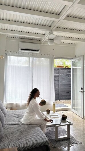 Ein Zimmer mit großem Fenster, Deckenventilator und einer Person, die an einem Tisch mit einem Laptop sitzt.