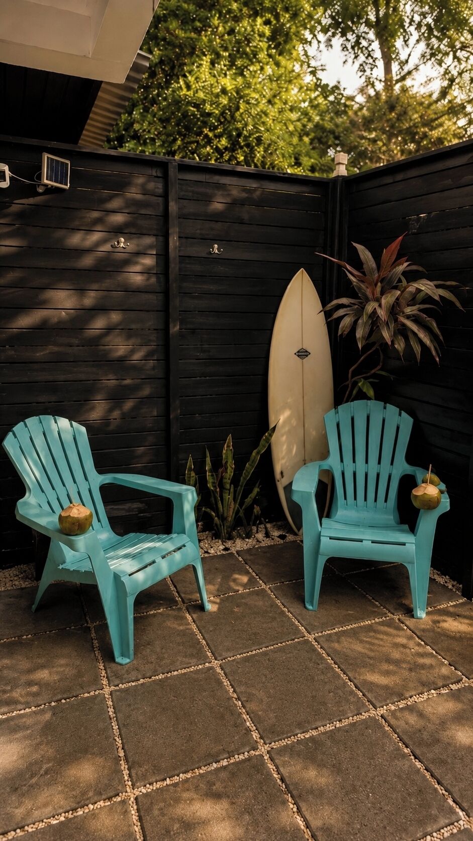 Two turquoise Adirondack chairs, a surfboard, and a potted plant in a shaded outdoor area with a dark wooden fence.