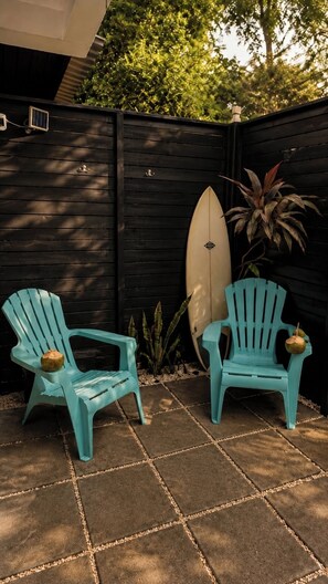 Dos sillas Adirondack turquesa, una tabla de surf y una planta en maceta en un espacio exterior sombreado con una cerca de madera oscura.