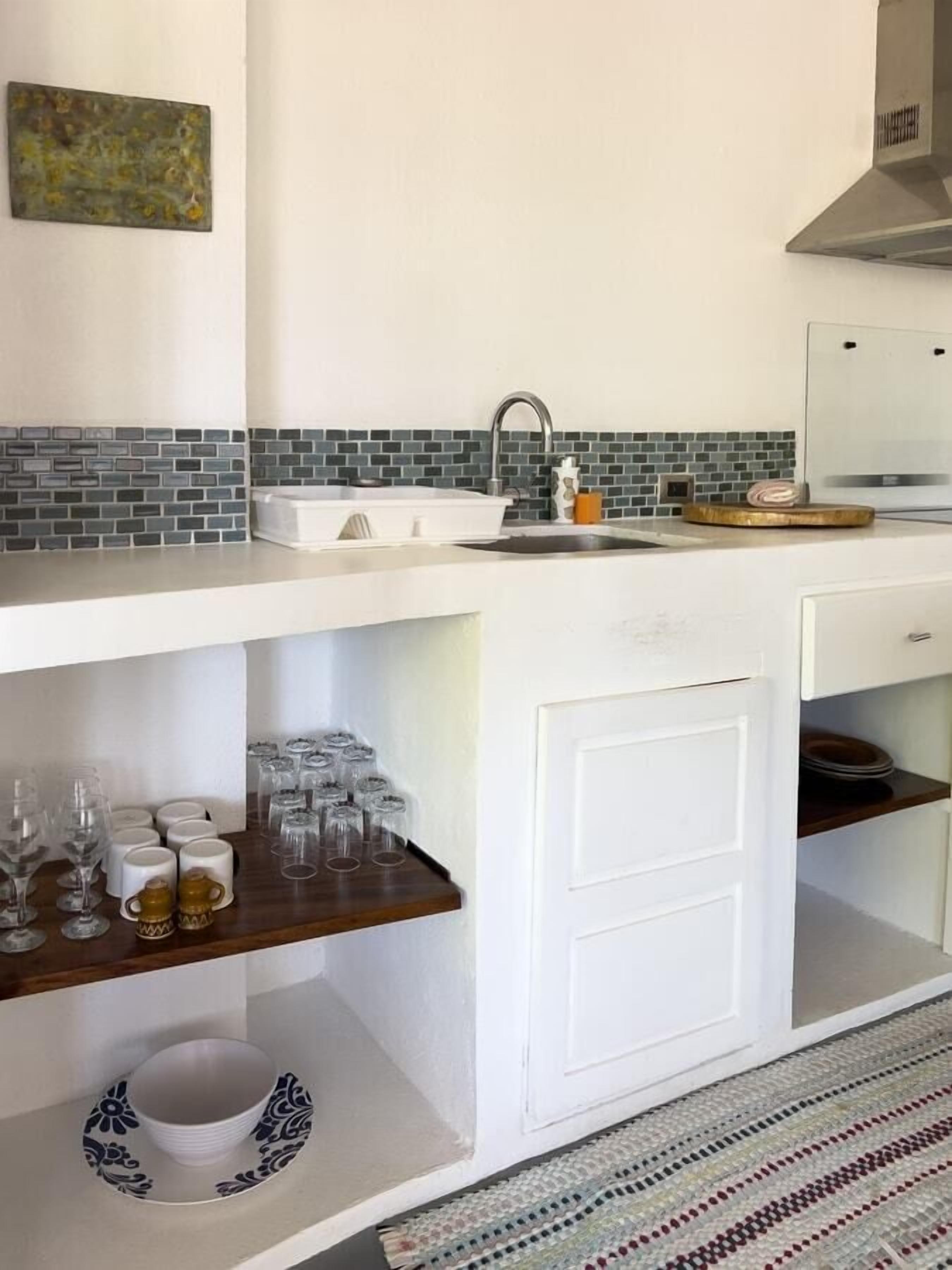 Une cuisine dotée d’un plan de travail blanc, d’un évier et d’un pare-feu composé de petites carreaux. On y trouve des étagères sous le plan de travail, où sont rangés divers objets en verre, ainsi qu’une armoire blanche avec une porte.