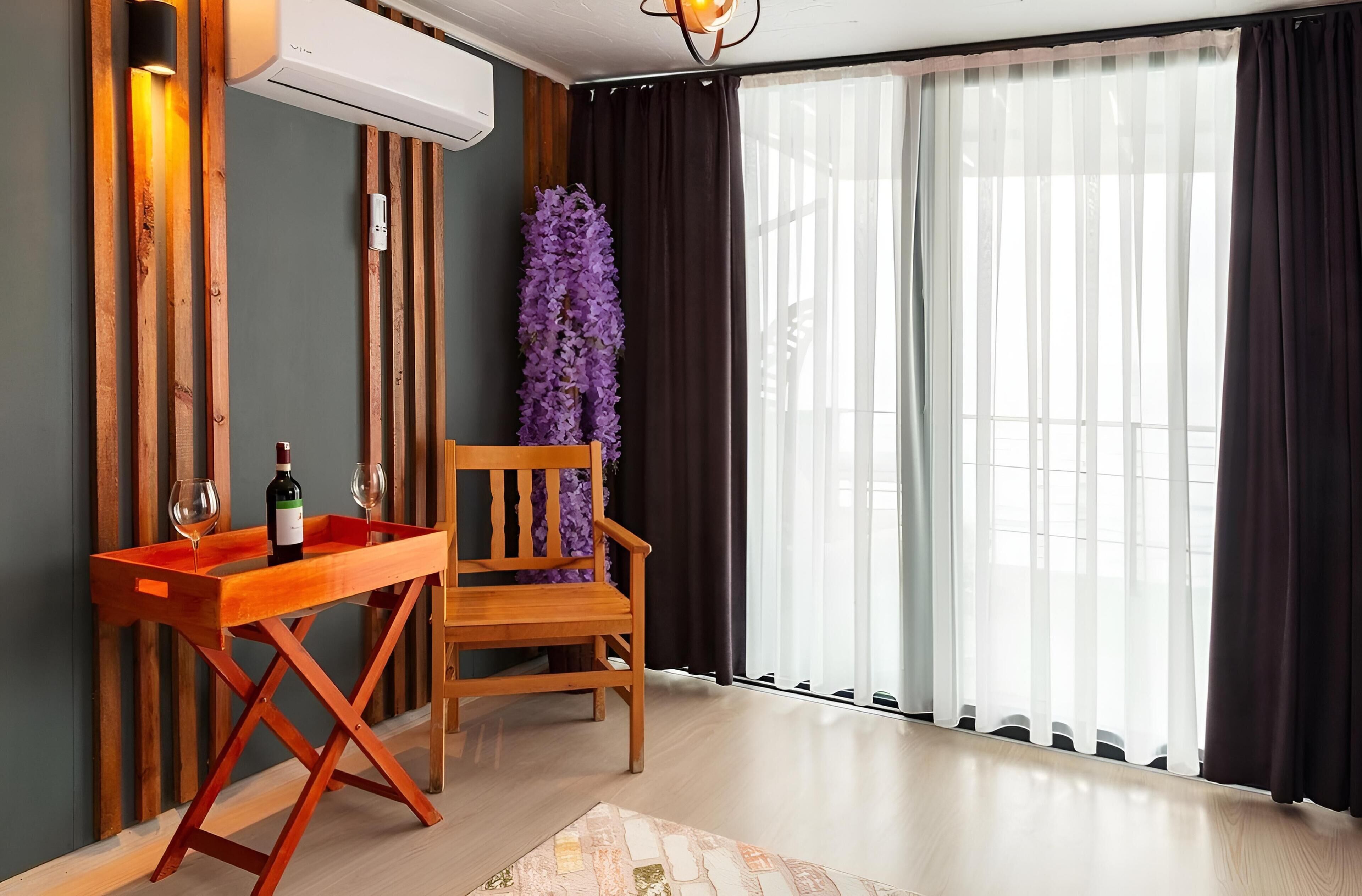 Une chambre moderne avec une chaise en bois, une petite table en bois sur laquelle se trouvent une bouteille et des verres, et une grande fenêtre ornée de rideaux légers.
