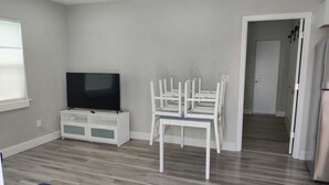 A modern living room with a flat-screen TV on a white entertainment unit, a dining area with white chairs, and a kitchen area with a refrigerator.