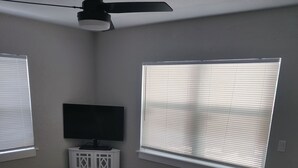 A bedroom with a bed, a TV on a cabinet, and two windows with blinds.