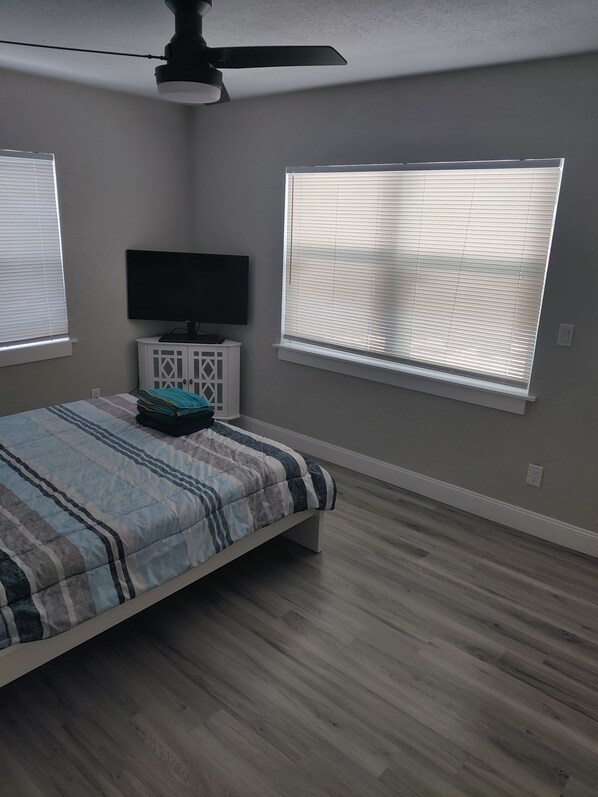A bedroom with a bed, a TV on a cabinet, and two windows with blinds.