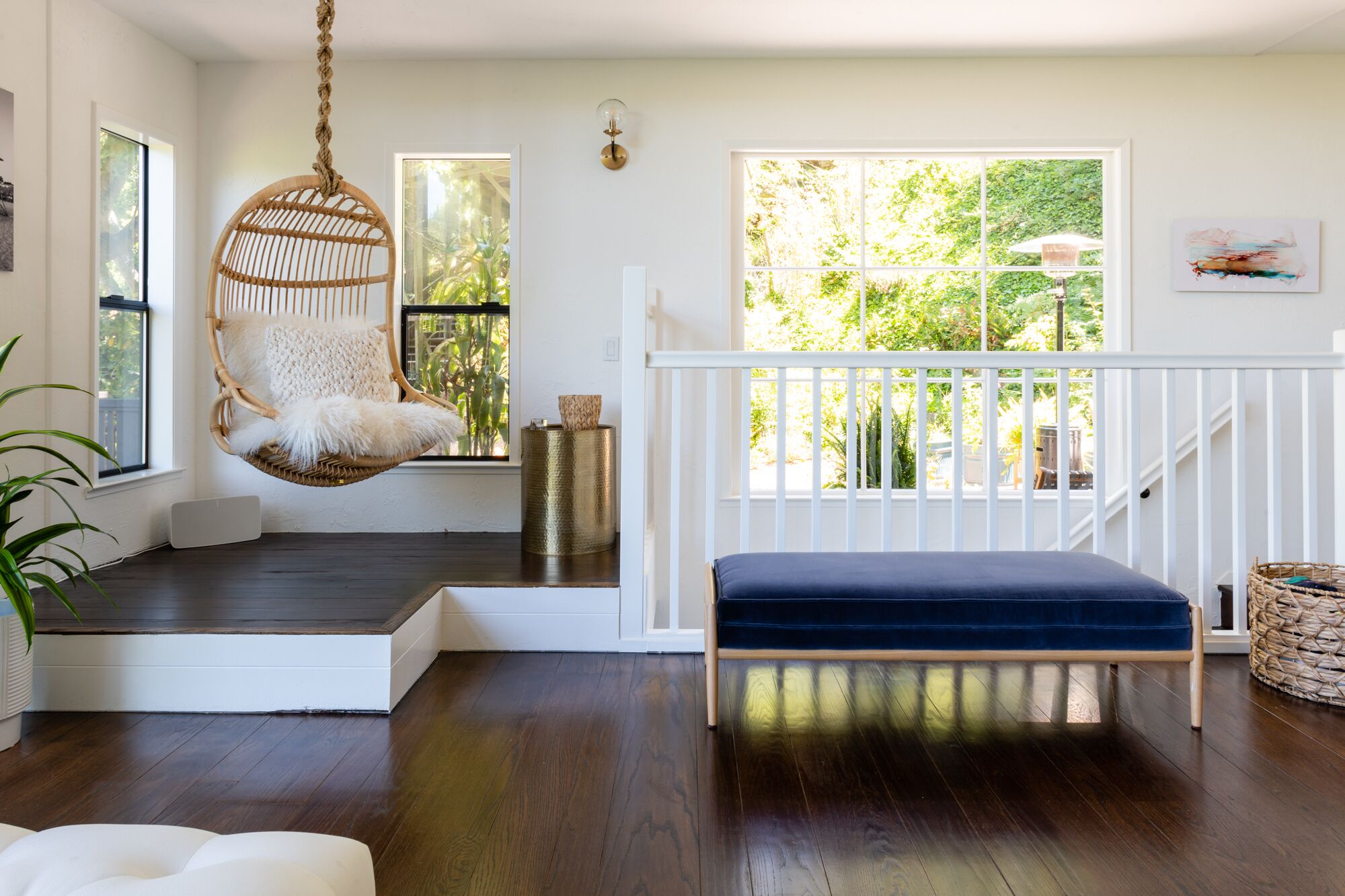 A room with a hanging chair, a blue bench, a white railing, and a view of greenery outside.
