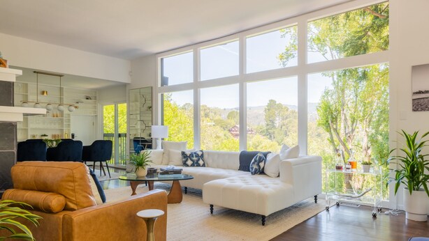 Un salon spacieux avec une grande fenêtre, un canapé blanc, un fauteuil en cuir marron et une table basse en verre.