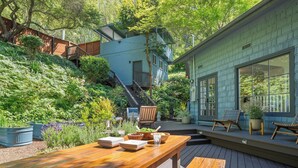 Une terrasse en bois avec des meubles d’extérieur, un escalier menant à une maison bleue et un jardin agrémenté de différentes plantes.