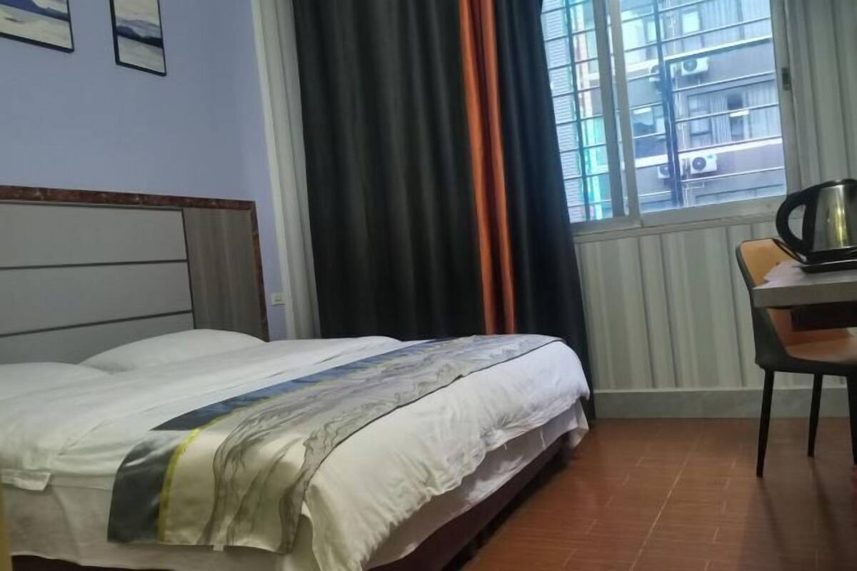A hotel room with a bed, a chair, a table with a kettle, and a window with curtains.