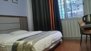 A hotel room with a bed, a chair, a table with a kettle, and a window with curtains.