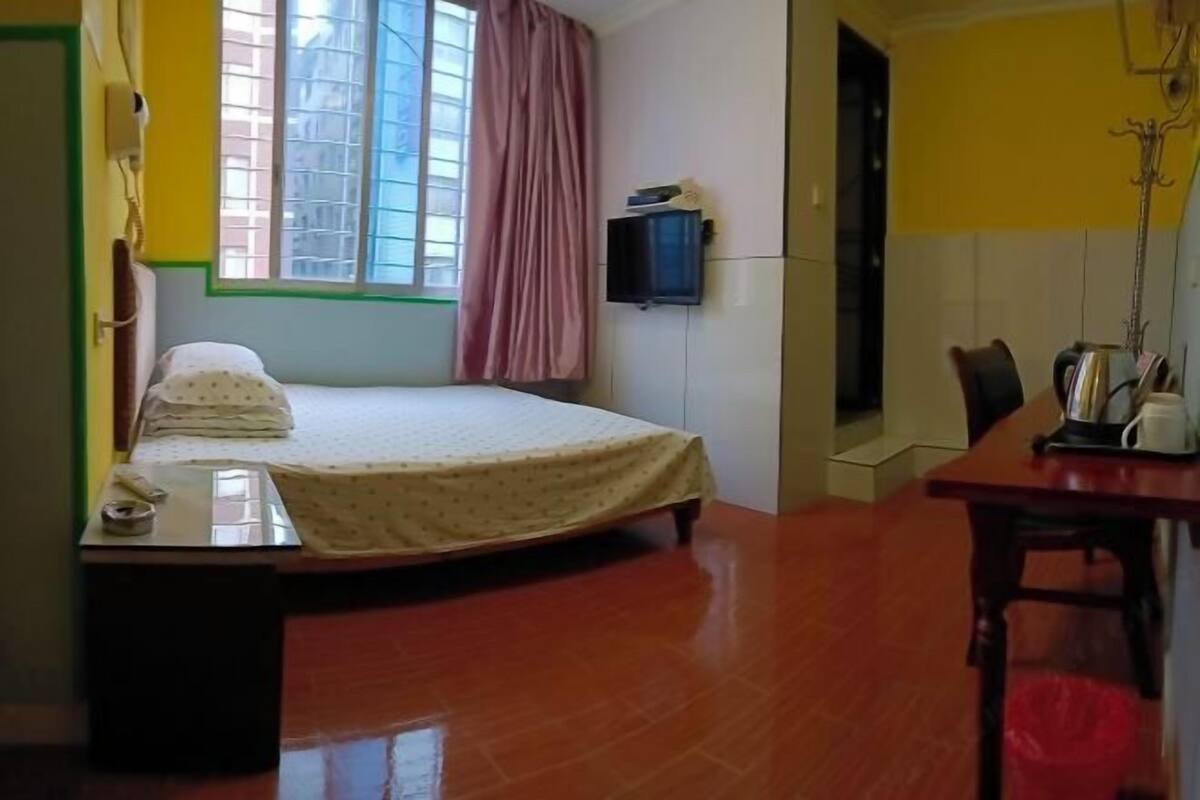 A room with a bed, a small table, a chair, a window with curtains, and a wall-mounted TV.