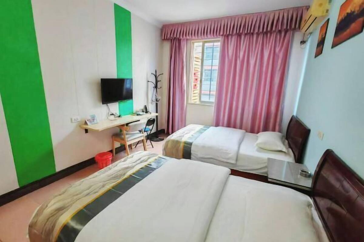 A hotel room with two beds, a desk with a television, and a window with pink curtains.