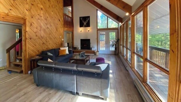 A spacious living room with a large couch, a fireplace, and large windows offering a view of the outdoors. - Beautiful cottage w/firepit, pool and a river in the property (Duclos)