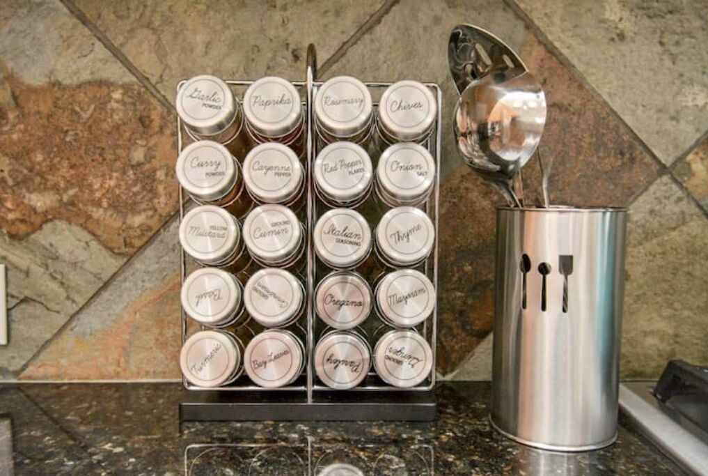 Un mueble de especias con varios frascos etiquetados y un recipiente metálico con una cuchara.