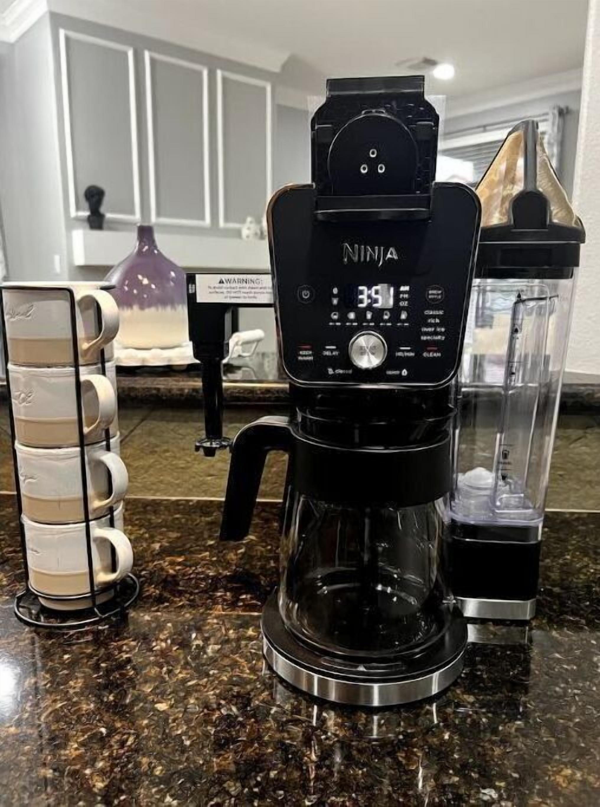 Eine Kaffeemaschine mit digitaler Anzeige, ein Stapel weißer Tassen und eine transparente Glascaraffe auf einer Granitabdeckung.