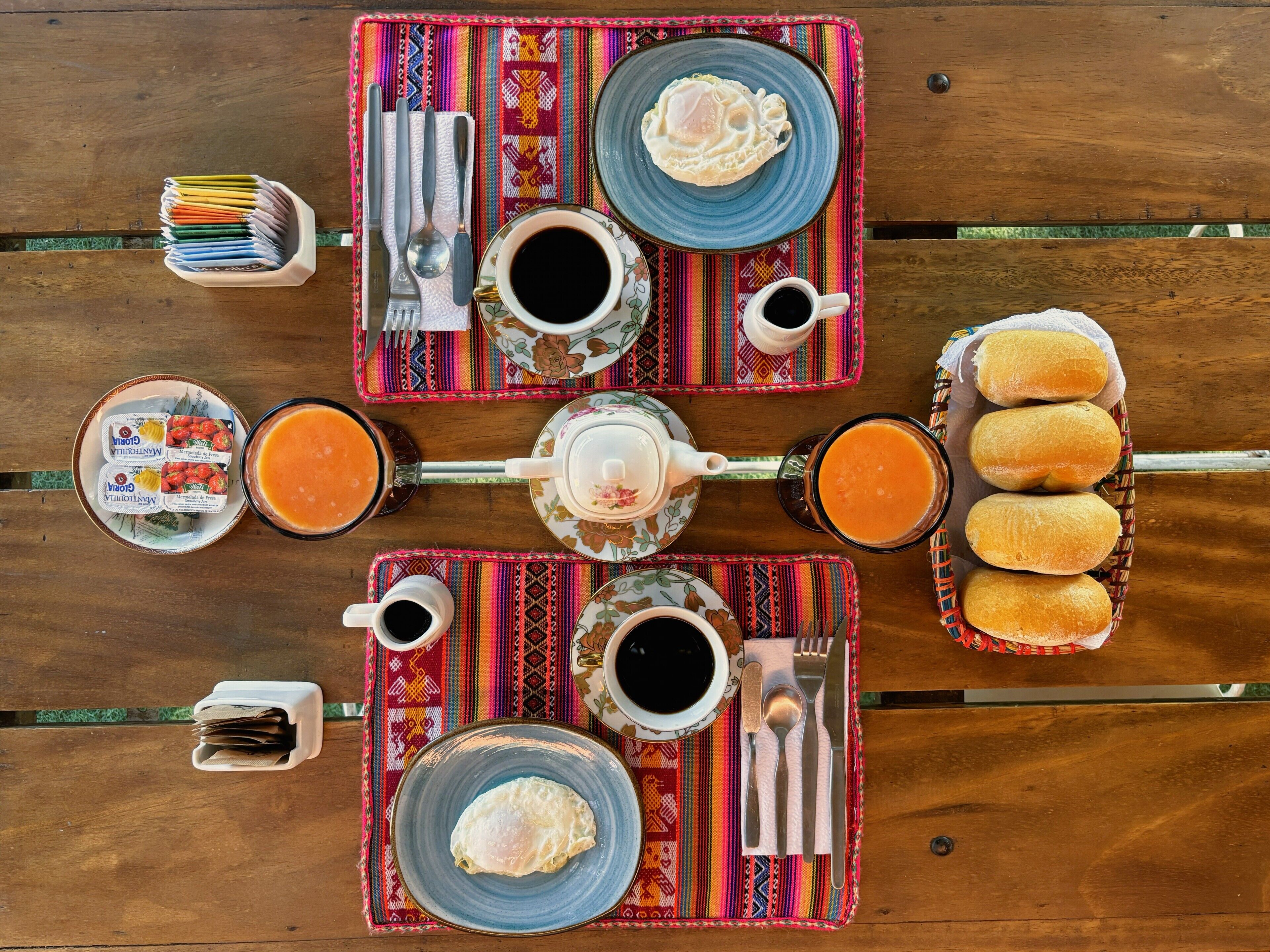A wooden table set with a meal including eggs, toast, and coffee, with colorful placemats and a variety of utensils.