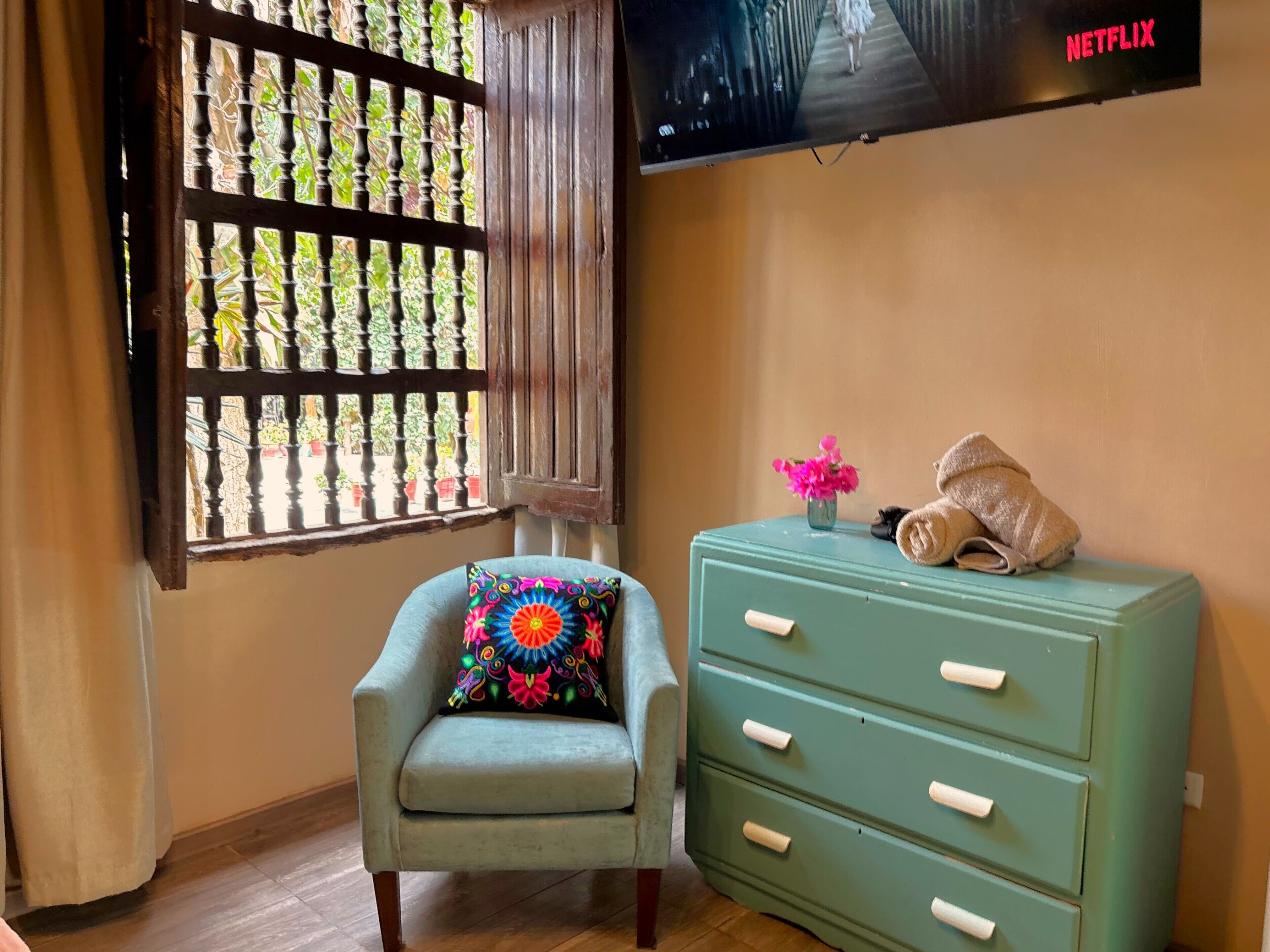 Una habitación con un sillón azul claro, un aparador verde con cajones, una ventana con celosía decorativa y un televisor mostrando el logotipo de Netflix.