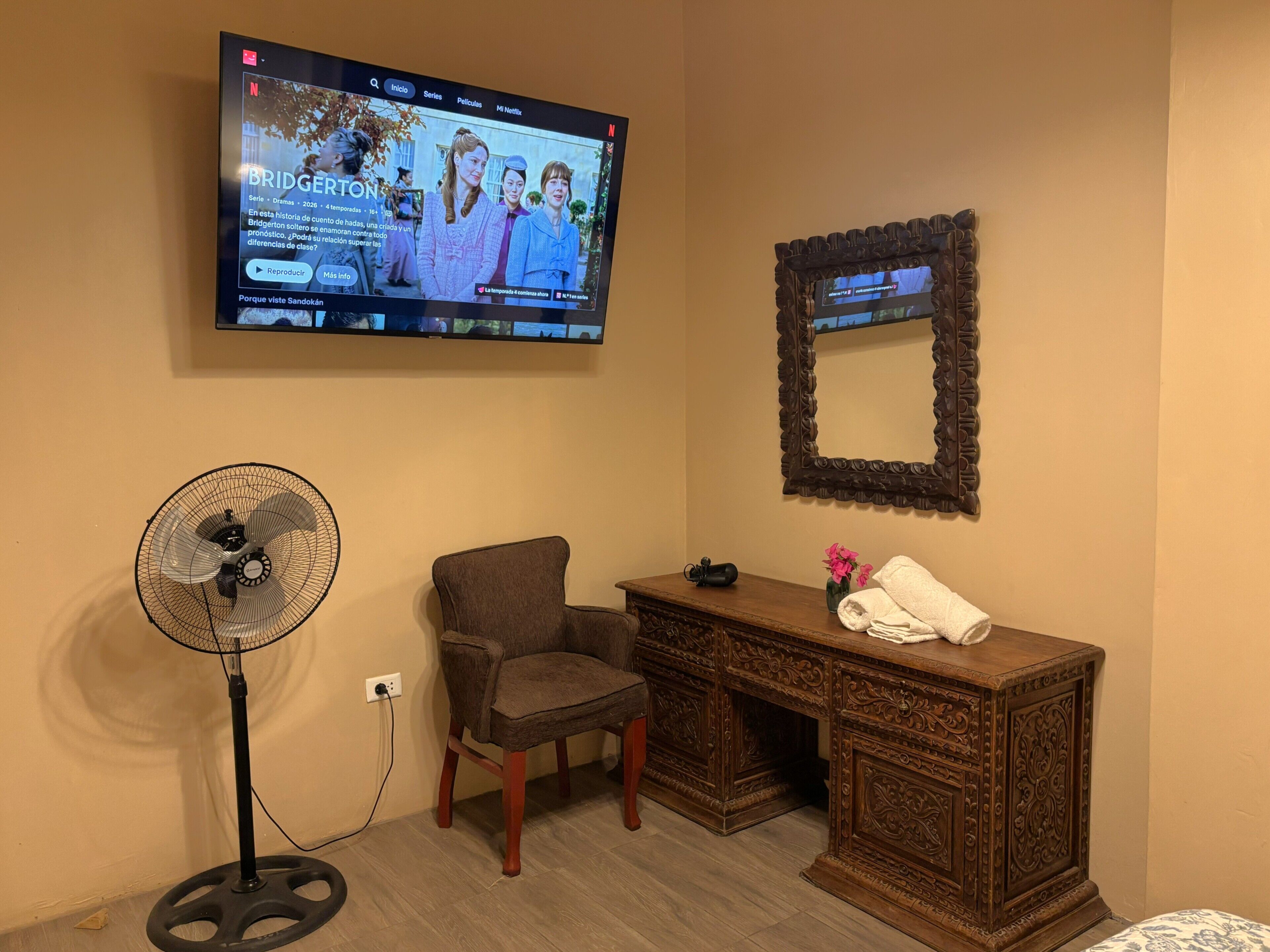 A room with a TV mounted on the wall, a standing fan, a wooden desk, and a chair.