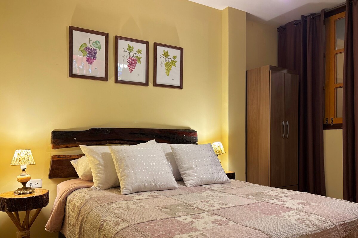 Quarto com cabeceira de madeira, uma cama grande com roupa de cama estampada, mesa de cabeceira com abajur e um guarda-roupa.
