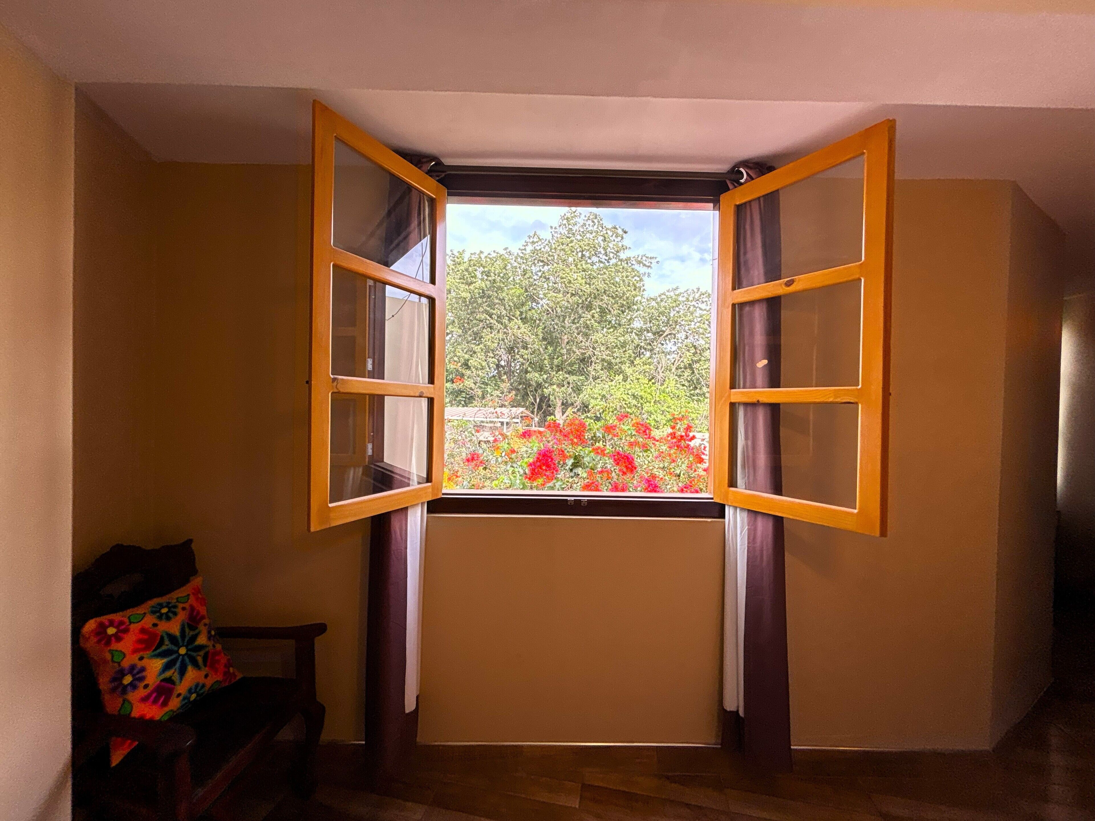 Una habitació amb una finestra oberta que mostra un jardí amb flors vermelles i arbres.
