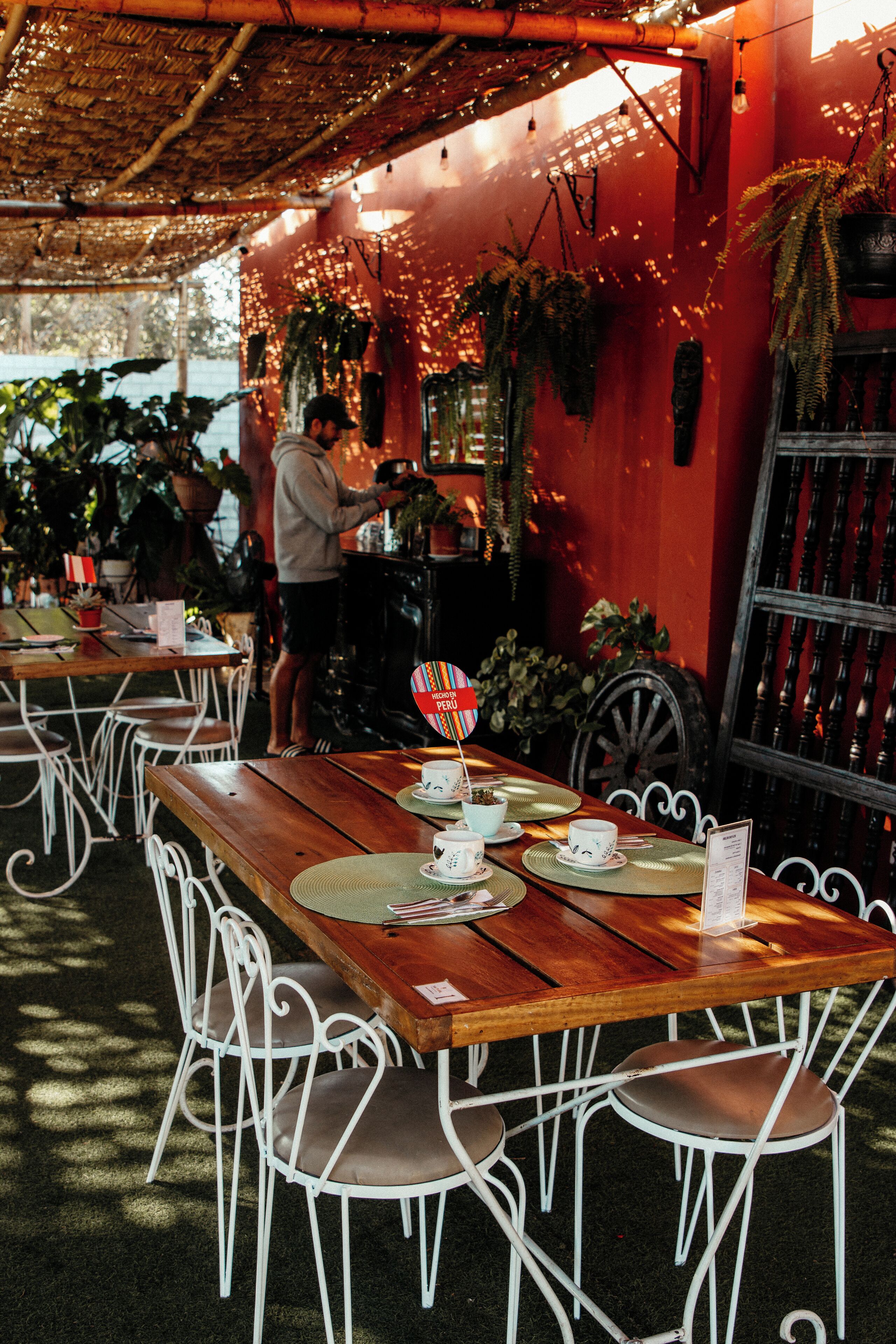 A restaurant with wooden tables and white metal chairs, a red wall, and hanging plants.