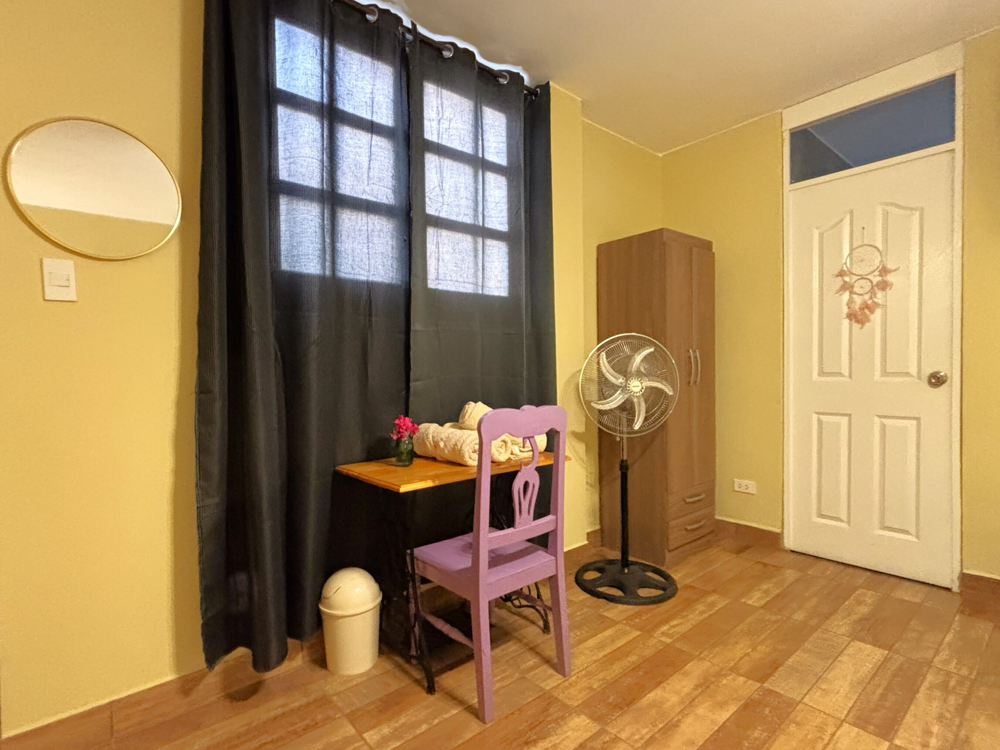 A room with a wooden floor, a black curtain, a wooden cabinet, a white door, a purple chair, a small table with a vase of flowers, and a standing fan.