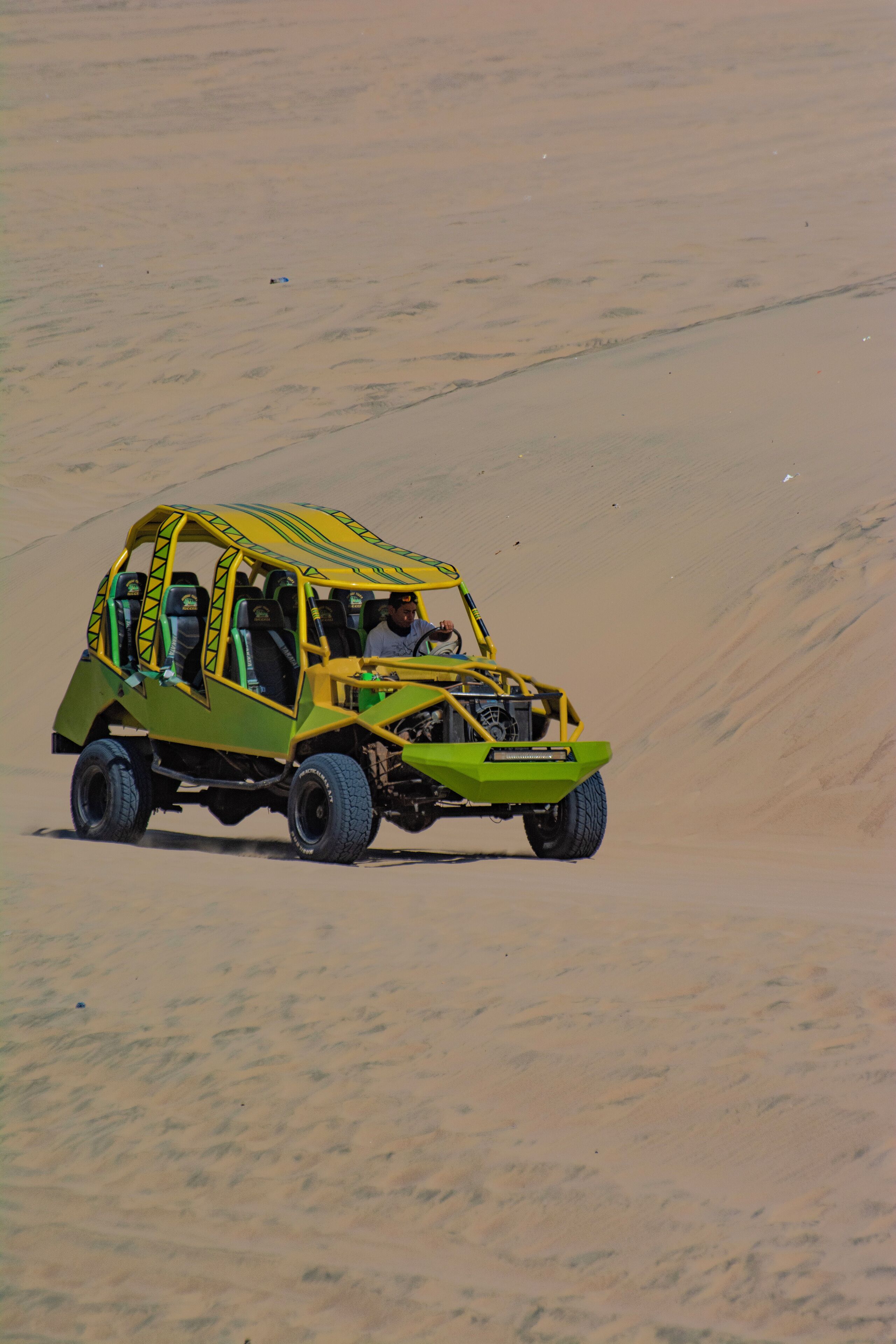 Un vehículo todoterreno en el desierto con carrocería verde y amarilla, circulando sobre dunas de arena.