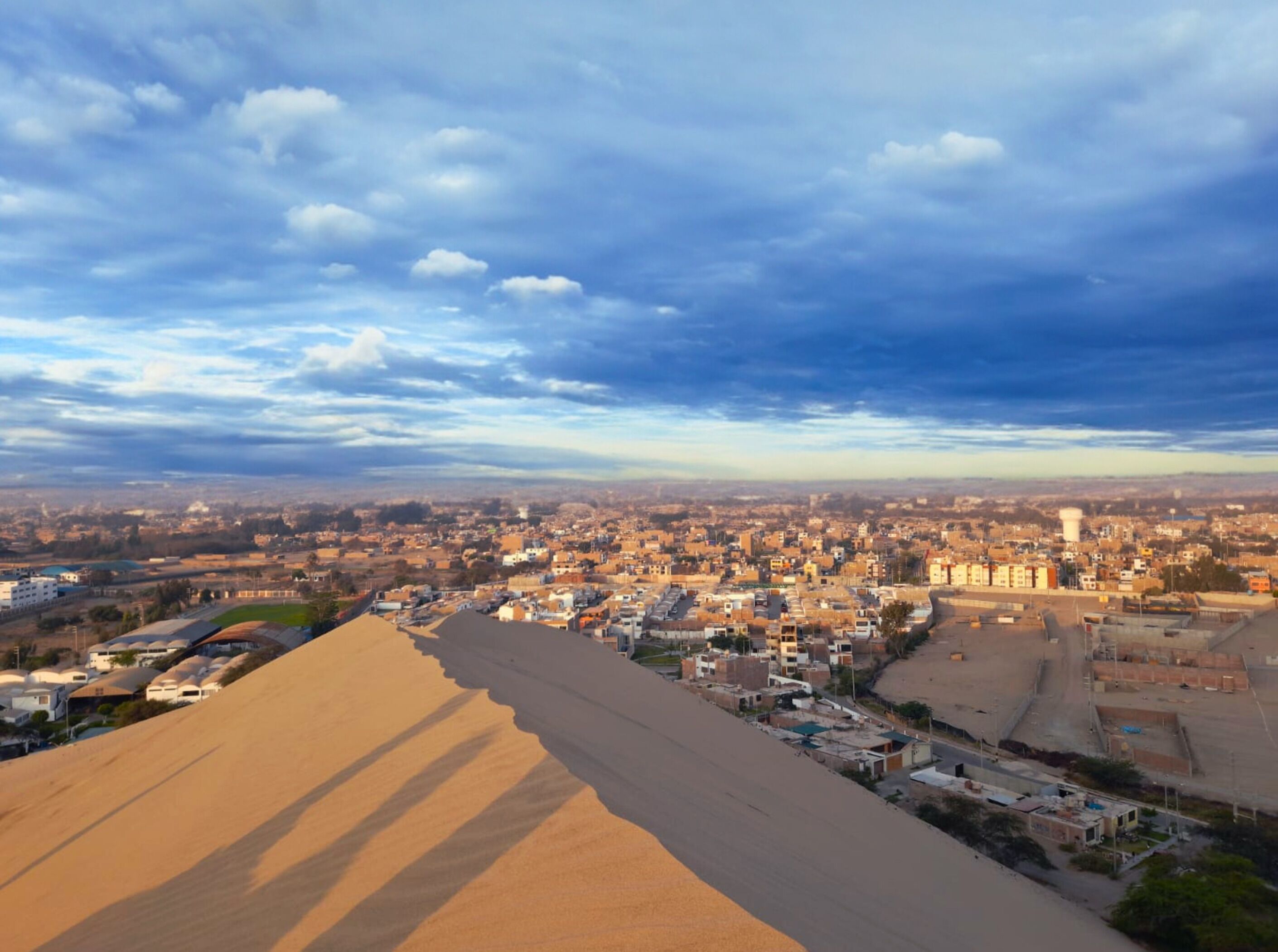 Un paisaje urbano con una duna de arena destacada en primer plano, edificios en el segundo plano y un cielo nublado al fondo.