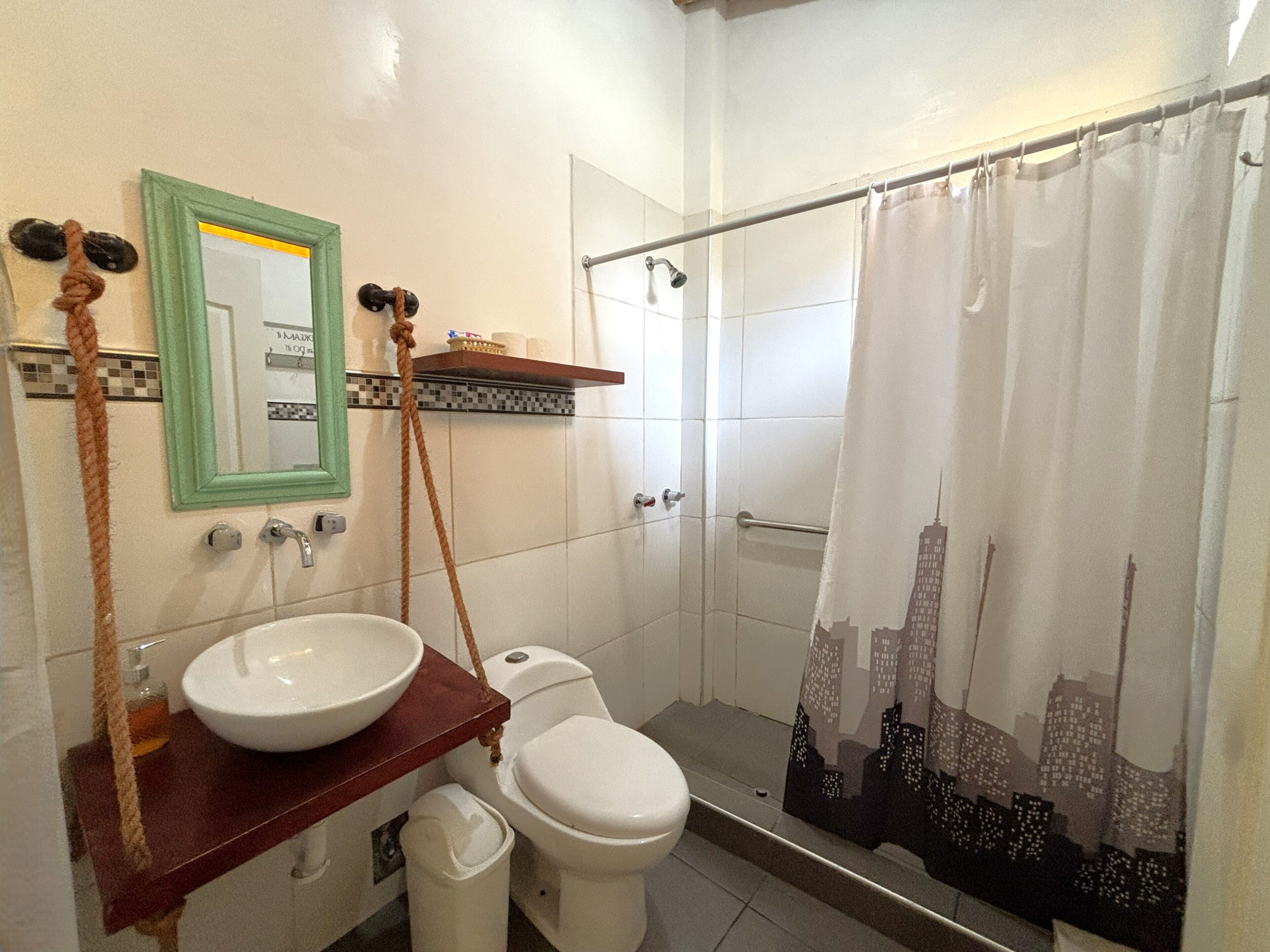 Baño con lavamanos, inodoro y ducha de color blanco. Se ve un espejo con marco verde y un perchero con cuerda.