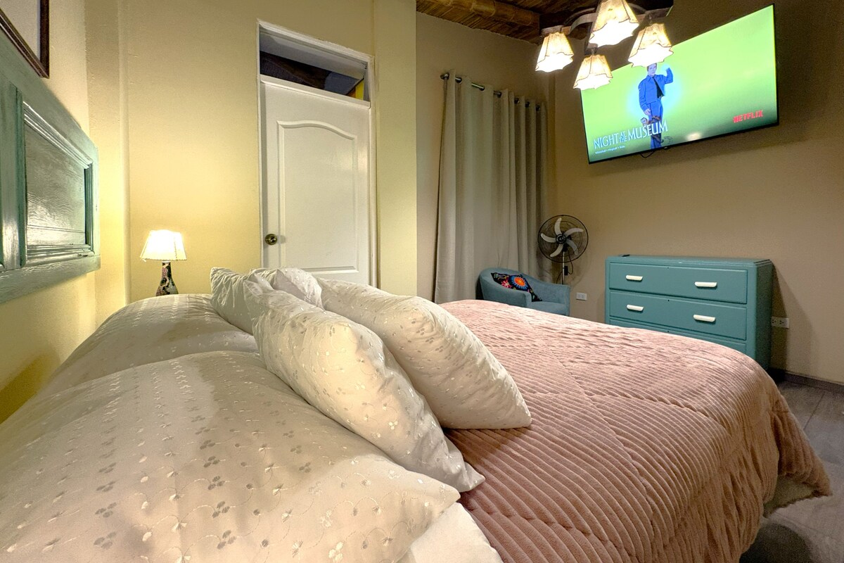 Quarto com cama, televisão na parede, ventilador, penteadeira e porta.