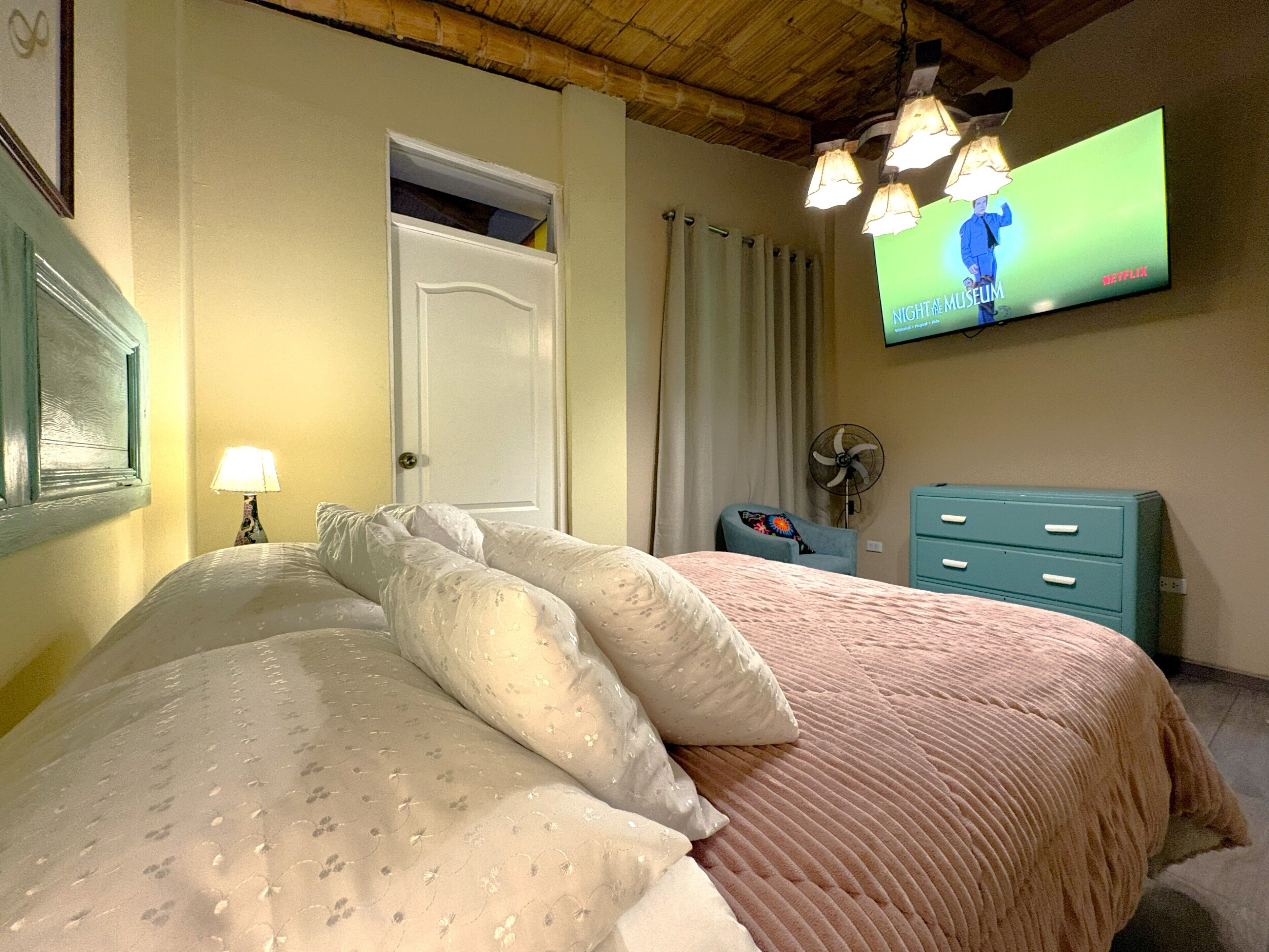 A bedroom with a bed, a TV mounted on the wall, a fan, a dresser, and a door.