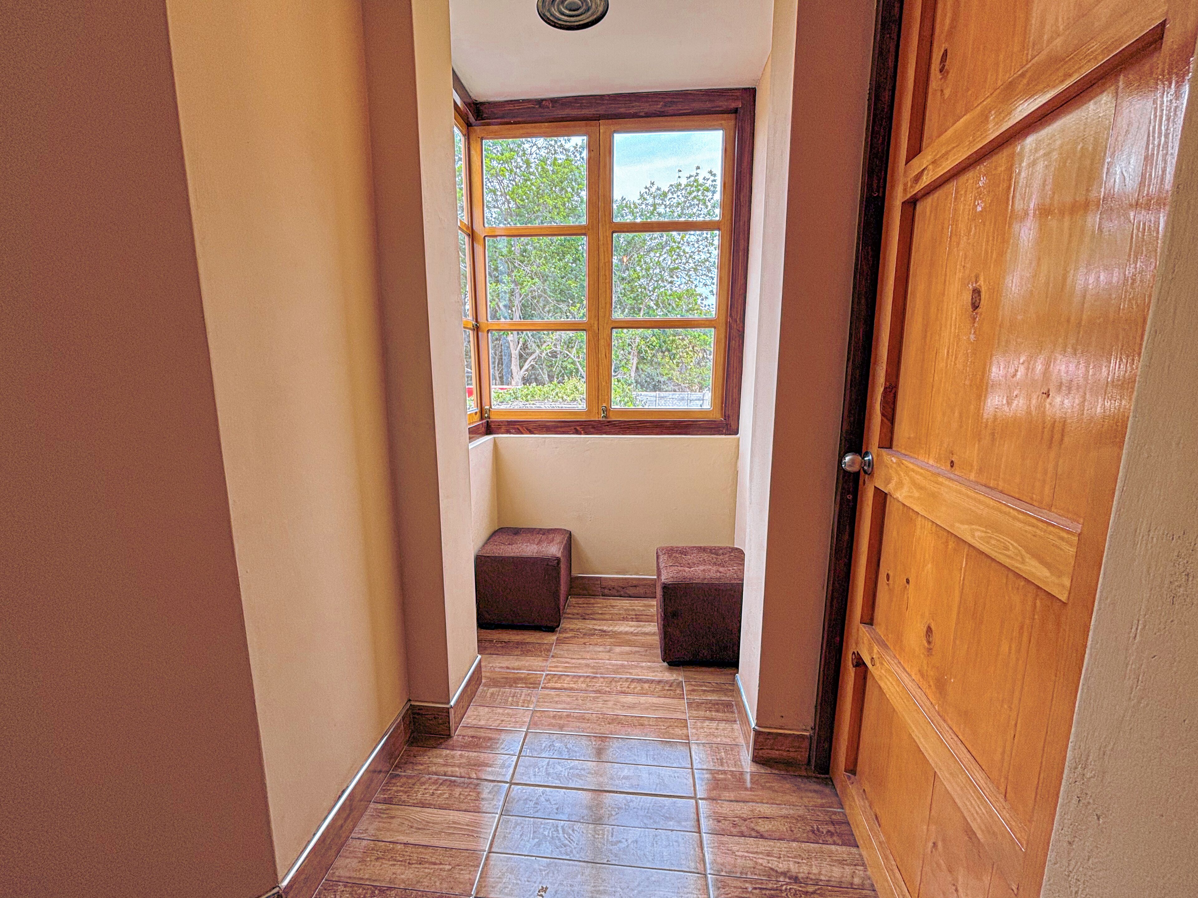 A narrow hallway with a wooden door on the right, a window at the end, and two small stools in the center.