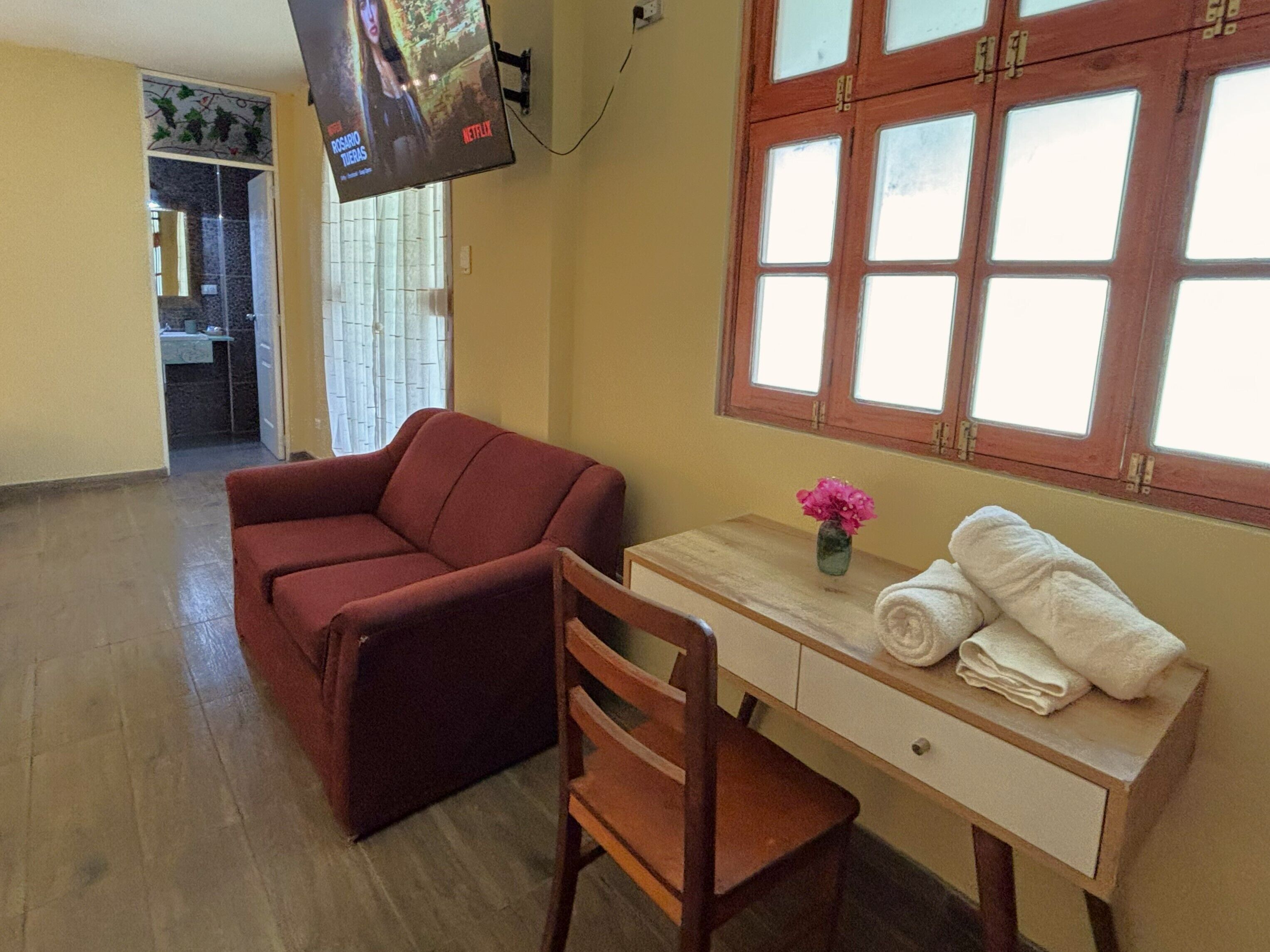 Una habitación con un sofá rojo, un escritorio de madera con un jarrón de flores y una ventana con cortinas.