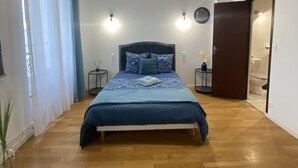 Une chambre avec un plancher en bois, un lit recouvert d’une courtepointe bleue, un luminaire suspendu et une porte donnant sur une salle de bain.