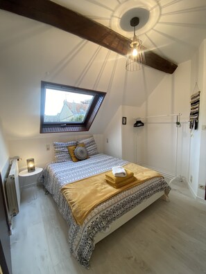 Un dormitorio con tragaluz, una cama con ropa de cama estampada, una mesita de noche con lámpara y una toalla colgada.