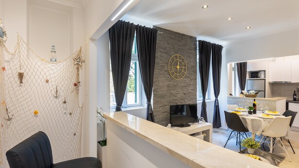 Een moderne keuken met een achterwand van steen, een eethoek met een witte tafel en stoelen, en een zithoek met een donkere bank en een decoratieve wand.
