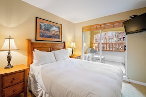 A bedroom with a bed, two bedside tables with lamps, a wooden headboard, a framed picture on the wall, and a balcony with a view.