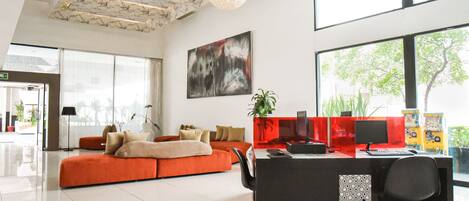 A modern hotel lobby with a large orange sofa, a black desk with multiple monitors, and a large abstract painting on the wall.