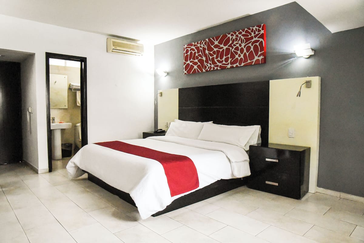 Une chambre d’hôtel moderne avec un grand lit, une table de chevet, une lampe fixée au mur et une salle de bain visible à travers une porte ouverte.