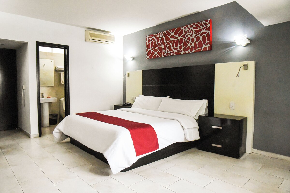 Una habitación de hotel moderna con una cama grande, una mesita de noche, una lámpara de pared y un baño visible a través de una puerta abierta.