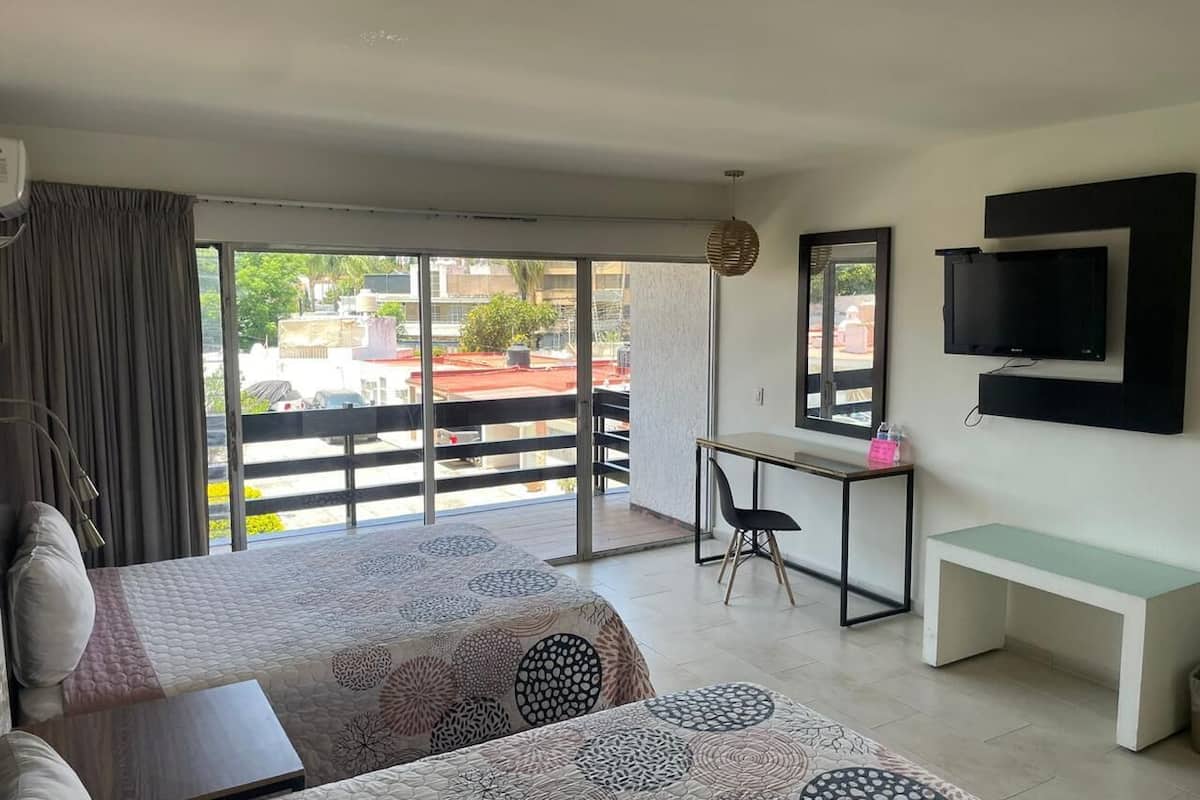 Une chambre d’hôtel avec deux lits, un bureau, une télévision et un balcon offrant une vue sur les immeubles et les arbres.