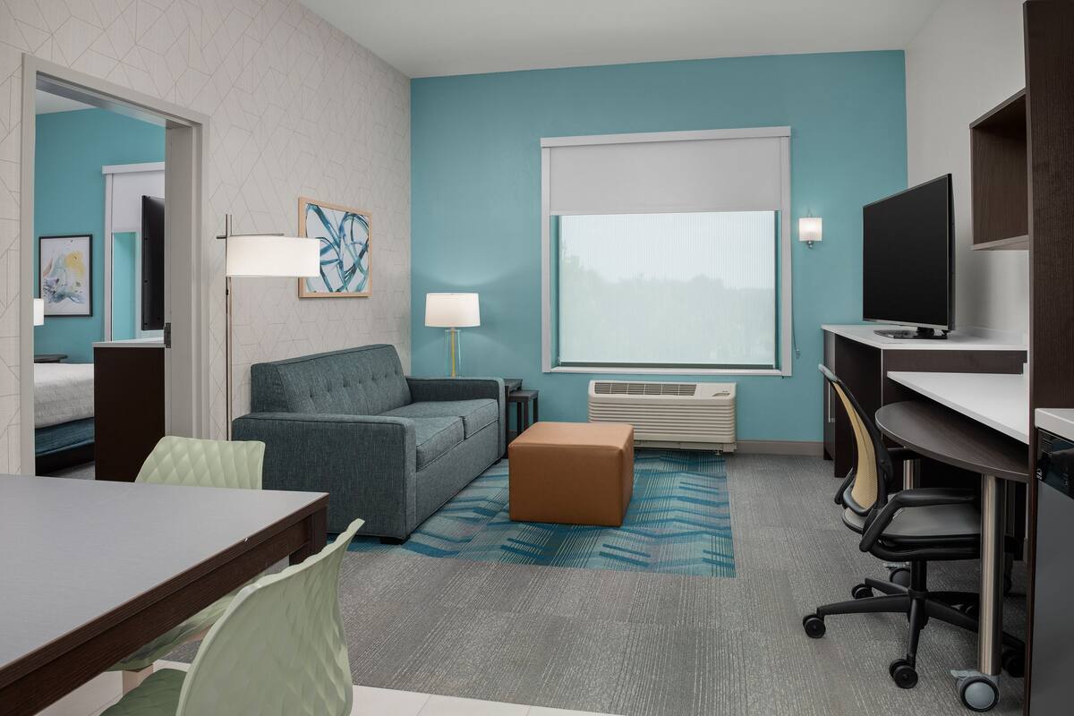 Een moderne hotelkamer met een grijze bank, een klein bruin poefje, een bureau met een computer, een televisie en een groot raam met uitzicht op de stad.