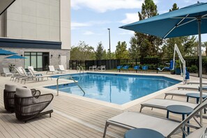 Ein Schwimmbecken mit Liegestühlen und Sonnenschirmen, umgeben von einer Holzterrasse und einem Zaun.