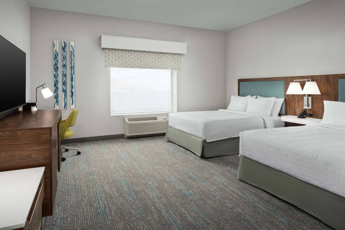 A hotel room with two beds, a desk, a chair, a TV, and a window with blinds.