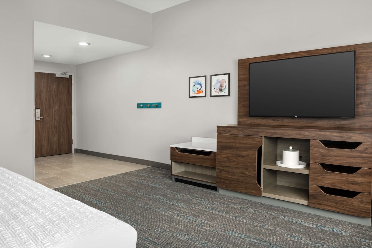 A modern hotel room with a large bed, a flat-screen TV mounted on a wooden cabinet, a white sink, and a small bathroom.